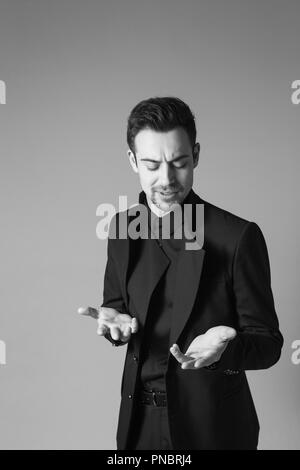 Close up portrait of a young handsome man in a black suit, standing, seriously looking at his hands while explaining something, against plain studio b Stock Photo