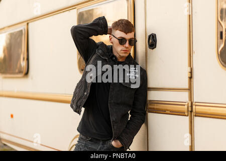 Fashionable young man in black stylish denim clothes posing near a vintage trailer Stock Photo