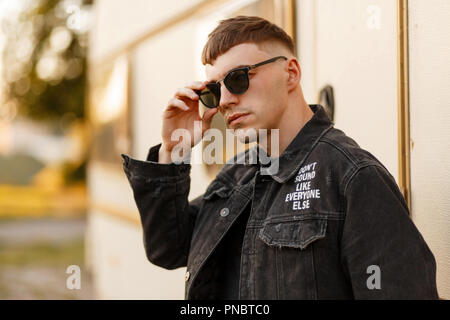 Stylish young man in black fashion glasses in black denim clothes posing near a vintage trailer Stock Photo