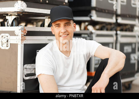 cheerful young happy man in a stylish black cap with a white long T-shirt sitting near containers on the street Stock Photo