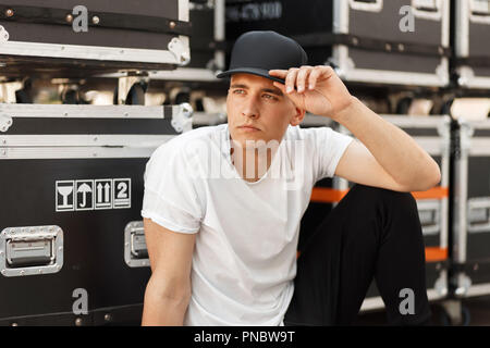 Beautiful model stylish man in a black cap and a white T-shirt sitting near containers on the street Stock Photo