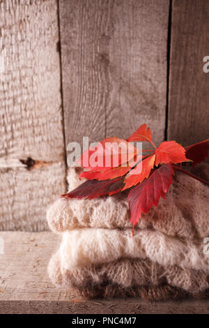 Stack of brown cozy knitted sweaters on a wooden table and red leaves, Cosy and soft warm autumn fall background, vertical Stock Photo