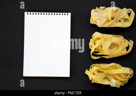 Uncooked pasta pappardelle and blank notepad on black background. From above. Copy space. Stock Photo