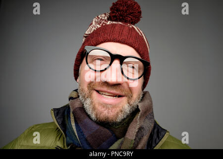 Portrait of mature hipster man in glasses and warm hat posing on gray background. Smiling bearded man wearing warm clothes for winter studio shot. Clo Stock Photo