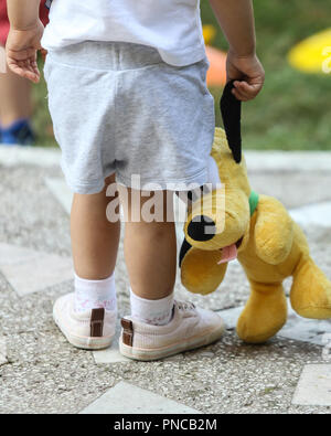 Young girl holding a plush toy in the front yard of a kindergarten while a man passes behind her Stock Photo