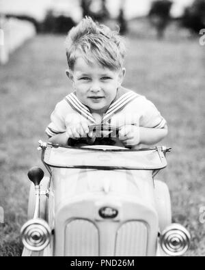 1930s BOY IN SAILOR SUIT DRIVING RACING TOY PEDDLE CAR LOOKING AT CAMERA HUGGING THE STEERING WHEEL WITH BOTH HANDS  - j6333 HAR001 HARS SPEED SAILOR HOME LIFE LUXURY AUTOMOBILE MALES CONFIDENCE TRANSPORTATION B&W EYE CONTACT HAPPINESS HEAD AND SHOULDERS ADVENTURE BOTH AUTOS EXCITEMENT STEERING WHEEL IN SPEEDING AUTOMOBILES PEDDLING STYLISH VEHICLES GRASPING JUVENILES BLACK AND WHITE CAUCASIAN ETHNICITY HAR001 OLD FASHIONED PEDDLE CAR Stock Photo