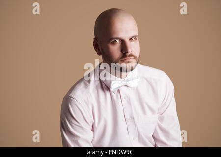 Portrait of handsome bearded bald man in light pink shirt and white bow, sitting on chair and looking at camera with serious face. indoor studio shot, Stock Photo