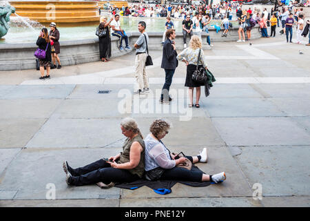 London England,UK,Trafalgar Square,public plaza,crowd,woman female women,senior seniors citizen citizens,sitting back to back on pavement ground,smart Stock Photo