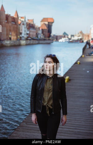 Female student wearing a leather jacket is walking by the river pier in Gdansk, Poland, smiling and looking down. Stock Photo