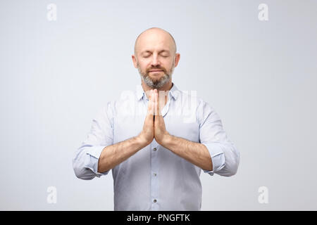 Yoga and meditation at office during work day. Handsome bald man with bristle keeping eyes closed while meditating, feeling relaxed, calm, peaceful ho Stock Photo