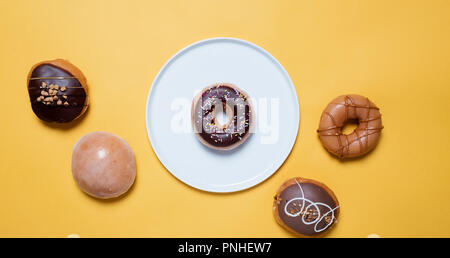Classic chocolate donuts with sprinkles on a white plate on a yellow background surrounded by various other decorated donuts Stock Photo