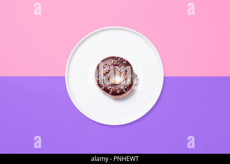 Classic ring donut with chocolate glaze frosting and sprinkles on a white plate on a split coloured pastel background of pink and purple. Stock Photo