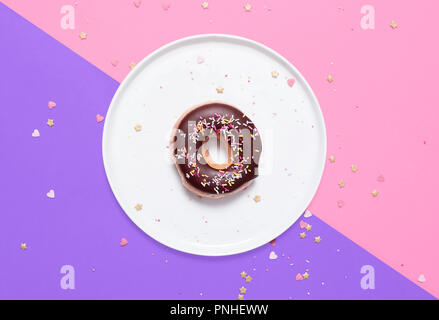 Classic donut with chocolate glaze frosting and sprinkles on a white plate on a split pink and purple pastel background with hearts scattered Stock Photo