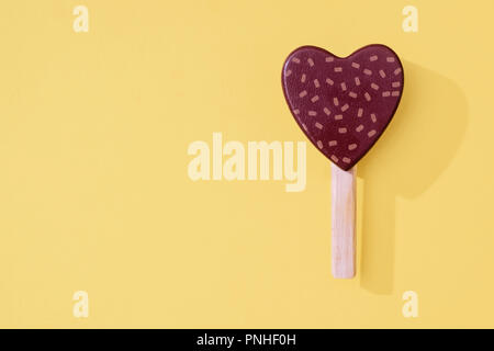 Wooden toy chocolate heart ice lollipop popsicle with sprinkles on a yellow background with wooden stick and space for copy and text. Stock Photo