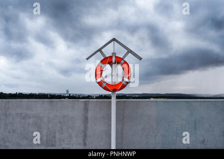 Orange lifebuoy on a stand on harbor pier against dramatic clouds in the sky. Stock Photo