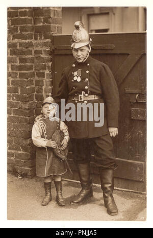 Original photographic postcard of fireman hero in uniform with medals posing outside for a photograph with his young son who is dressing up as a fireman with fireman's uniform, holding a fire hose, circa 1910, Manchester U.K. Stock Photo