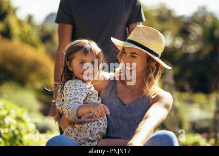 Cute little girl with her mother sitting on wheelbarrow being pushed by a man. Young family having fun outdoors in their farm. Stock Photo