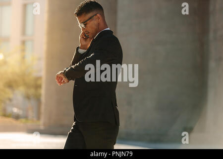 Man in formal clothes looking at his wrist watch and talking over mobile phone while walking on street. Busy office going person walking on street loo Stock Photo
