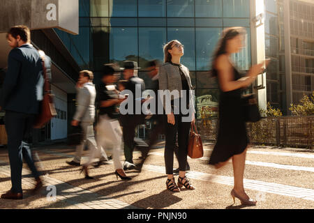 Businesswoman holding her hand bag standing still on a busy street with people walking past her using mobile phones. Woman standing amidst a busy offi Stock Photo