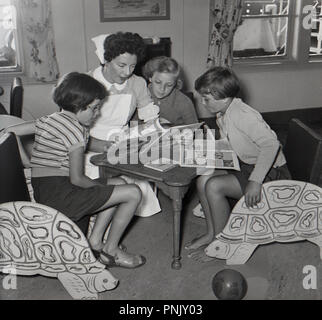 1950s, historical, in the nursery or playroom on a steamship heading from the Cape in South Africa, a female uniformed nurse sits with three young girls looking at the pages in a picture book. Stock Photo