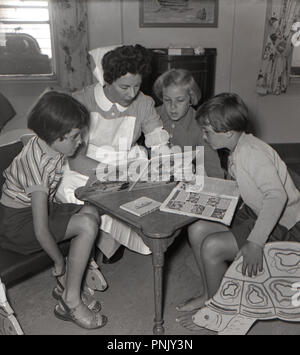 1950s, historical, in the nursery or playroom on a steamship heading from the Cape in South Africa, a female uniformed nurse sits with three young girls looking at the pages in a picture book. Stock Photo