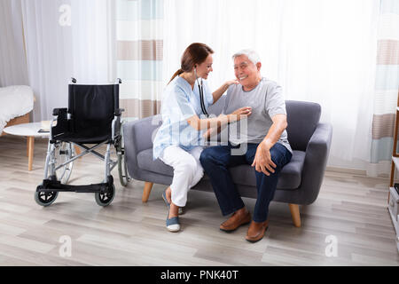 Female Nurse Checking Happy Senior Man Sitting On Sofa With Stethoscope Stock Photo