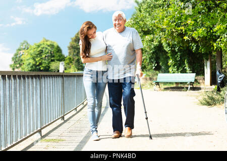 Young Woman Assisting Her Happy Father While Walking Near Railing In Park Stock Photo