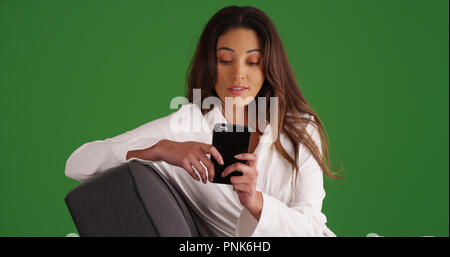Attractive Latina in white bathrobe on couch using smartphone on green screen Stock Photo