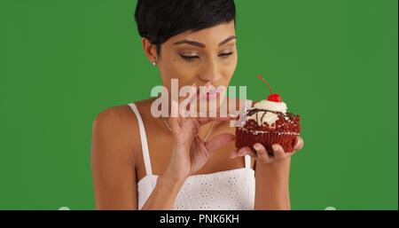 Young black woman enjoying cupcake with icing and cherry on top on green screen Stock Photo