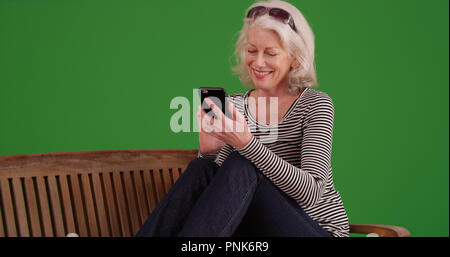 Smiling old white female using mobile device on park bench on green screen Stock Photo