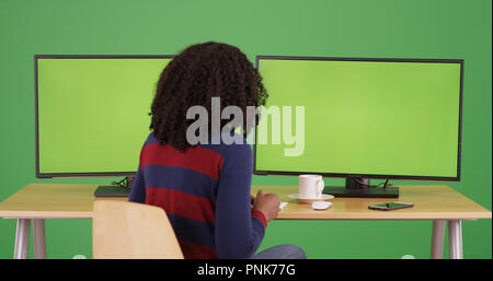 Black woman sitting by computers with green screens on display on green screen. Stock Photo