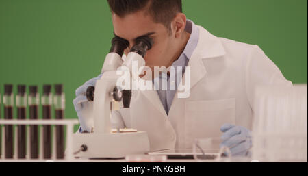 Millennial Hispanic medical scientist looking through microscope on green screen Stock Photo