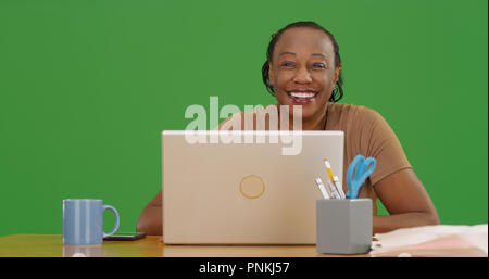 Old black woman sitting at desk with laptop smiling at camera on green screen Stock Photo