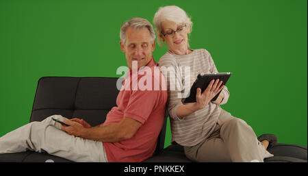 Senior couple sitting on couch using technology on green screen Stock Photo