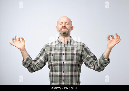 Yoga and meditation concept. Handsome bald man with bristle keeping eyes closed while meditating, feeling relaxed, calm, peaceful holding hands in mud Stock Photo