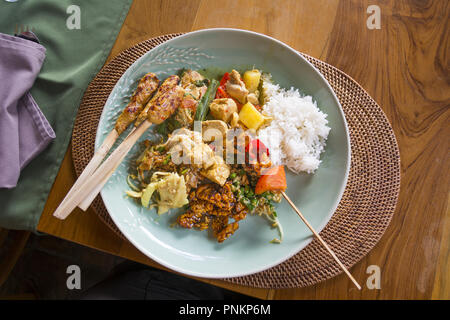 Plate of typical Balinese, Thai, Indonesian food with chicken satay sticks, rice, deep fried tempeh, steamed tuna,coconut and snake bean salad, vegeta Stock Photo