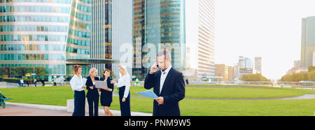 Businessman speaking by smartphone outside, keeping blue document case with employees in background. Concept of boss conversation by mobile phone in L Stock Photo
