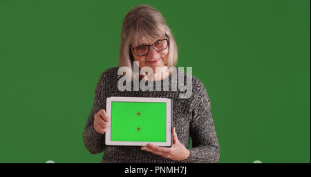 Smiling old woman showing tablet with green screen standing on green screen Stock Photo