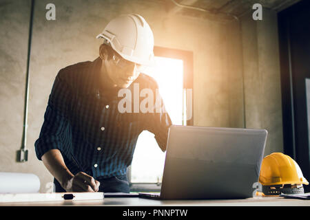 Man working, engineering checking, writing on blueprint and looking on laptop at site construction work. Stock Photo
