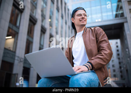Happy handsome student looking at computer screen while typing something on laptop keyboard during work break outdoors, teenager looking at the notebo Stock Photo