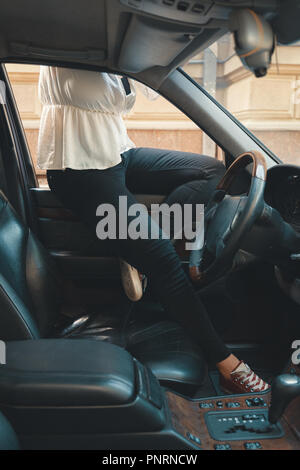 A cropped image of a woman sitting on the car window with crossed legs. Vehicle details, steering wheel, leather armchairs, gearbox. Stock Photo