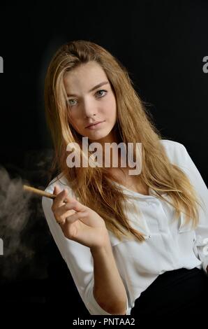 Young female sits with cigarillo and glass of whiskey in her hand. Elegant woman with white top and black dress. Stock Photo