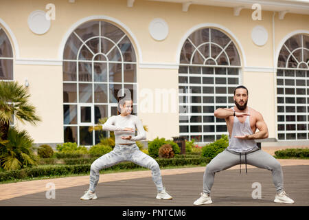 Sporty couple are stretching together on top of high urban building. They are sitting on mats and bending to straight less. They are finishing strength training with dumbbells Stock Photo