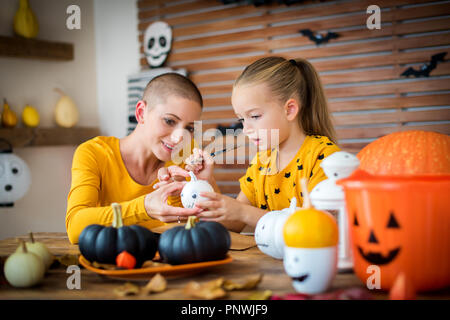 Cute young girl sitting at a table, decorating little white pumpkins with her mother, a cancer patient. DIY Halloween holiday and family lifestyle bac Stock Photo