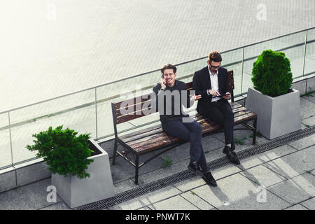 Two Businessman's have a conversation on street bench busy by phone and tablet. Stock Photo
