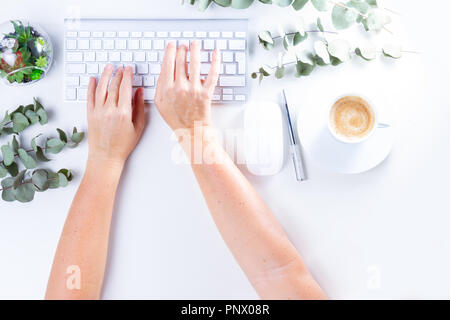 Flat lay home office workspace, someones two hands typing on white modern keyboard and silver green twigs Stock Photo