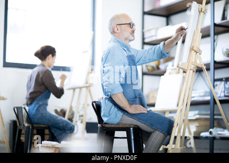 Mature man in workwear sitting in front of easel and sketching on paper sheet before painting Stock Photo