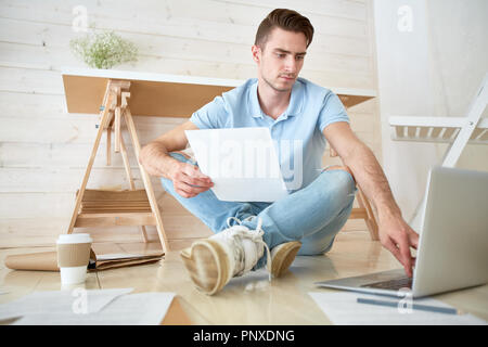 Young economist with paper sitting on the floor and searching for online data in laptop Stock Photo