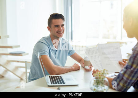 Smiling young businessman with drink looking at his colleague during discussion of papers Stock Photo