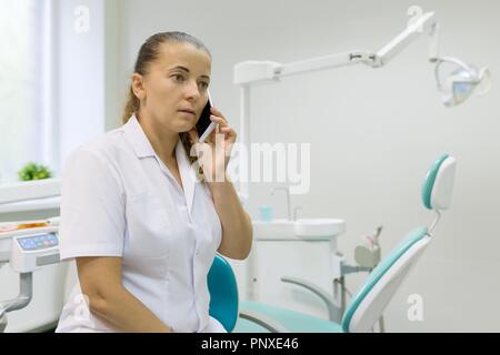 Portrait of a female dentist, doctor talking on mobile phone on dental chair background. Medicine, dentistry and healthcare concept. Stock Photo